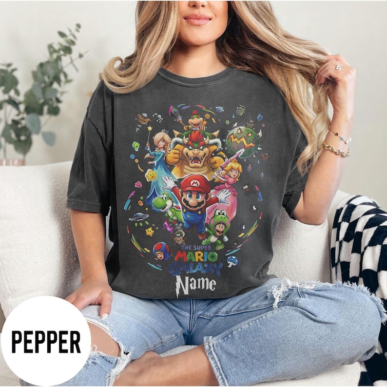 Puede incluir: Camiseta gris oscuro con un gr&aacute;fico colorido de Mario y otros personajes del juego Super Mario Galaxy. El dise&ntilde;o incluye el texto "THE SUPER MARIO GALAXY Name". La camiseta tiene una etiqueta Pepper.
