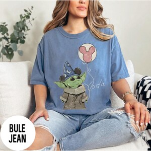K&ouml;nnte beinhalten: Hellblaues T-Shirt mit einer gr&uuml;nen Figur, die einen Zaubererhut mit Mickey Mouse-Ohren tr&auml;gt. Die Figur h&auml;lt einen Ballon mit einem Mickey Mouse-Kopf-Design. Das Wort "Yoda" ist darunter geschrieben.