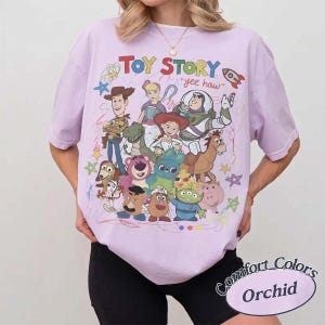 May include: An orchid-colored t-shirt featuring a graphic print of Toy Story characters including Woody, Buzz Lightyear, Jessie, and others. The text "TOY STORY" and "yee haw" appear above the characters, surrounded by colorful doodles of stars.