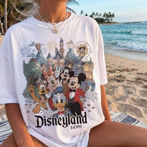 May include: A white short-sleeve t-shirt featuring a colorful graphic print of Mickey Mouse, Minnie Mouse, Donald Duck, Goofy, and Pluto in front of a castle and ferris wheel. The text Disneyland Est. 1955 is printed in black at the bottom.