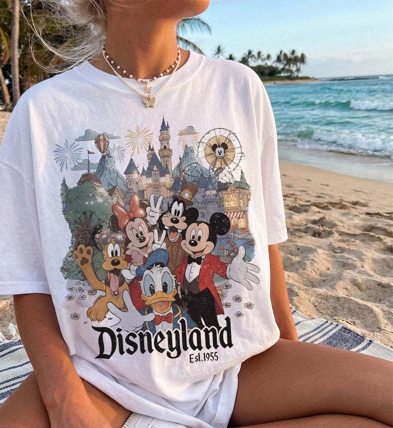May include: A white short-sleeve t-shirt featuring a colorful graphic print of Mickey Mouse, Minnie Mouse, Donald Duck, Goofy, and Pluto in front of a castle and ferris wheel. The text Disneyland Est. 1955 is printed in black at the bottom.