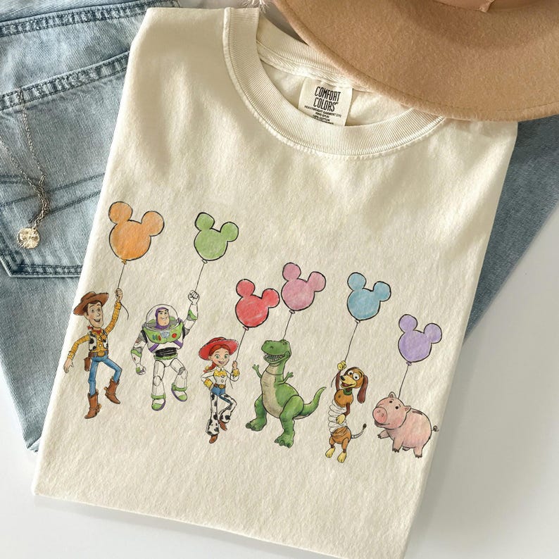 K&ouml;nnte beinhalten: Cremefarbenes T-Shirt mit einer farbenfrohen Illustration von Toy Story-Charakteren, die Mickey Mouse-f&ouml;rmige Ballons halten. Das Design umfasst Woody, Buzz Lightyear, Jessie, Rex, Slinky Dog und Hamm. Das Shirt liegt auf einer Jeans.