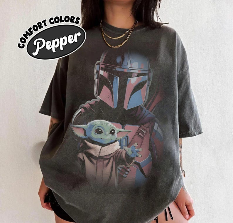 May include: A charcoal gray short-sleeve t-shirt featuring a graphic print of a helmeted warrior and a small green creature with large ears. The text COMFORT COLORS Pepper is printed in a black oval badge in the upper left corner of the image.