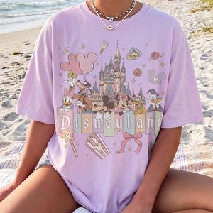 May include: A light purple short-sleeve t-shirt featuring a graphic print of the Disneyland castle, Mickey Mouse, Minnie Mouse, Goofy, Donald Duck, and Daisy Duck. The design includes pastel-coloured balloons, popcorn, and a bow with the text Disneyland.