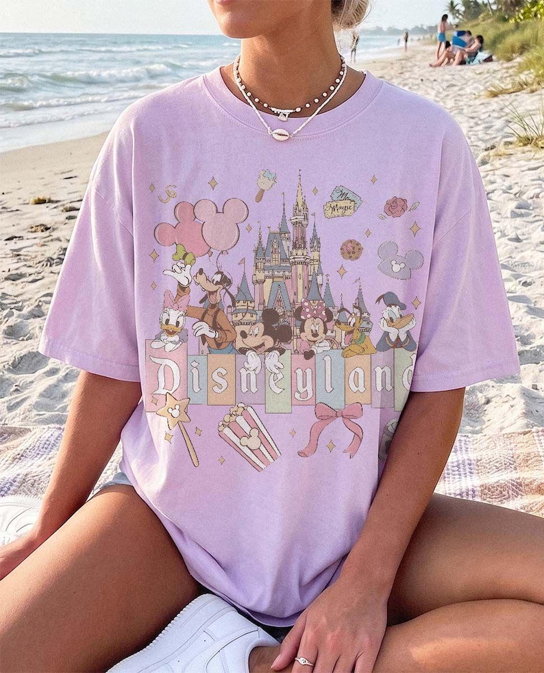 May include: A light purple short-sleeve t-shirt featuring a graphic print of the Disneyland castle, Mickey Mouse, Minnie Mouse, Goofy, Donald Duck, and Daisy Duck. The design includes pastel-coloured balloons, popcorn, and a bow with the text Disneyland.