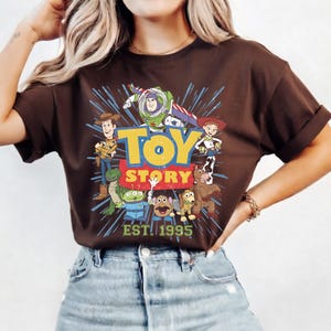 以下が含まれることがあります： 茶色のTシャツ。アニメ映画「トイ・ストーリー」のキャラクターのカラフルなグラフィックが特徴です。デザインにはウッディ、バズ・ライトイヤー、ジェシーなどのキャラクターと「TOY STORY」と「EST. 1995」の文字が含まれています。