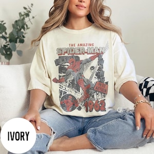 Pu&ograve; includere: T-shirt color avorio con grafica vintage di Spider-Man. Il design include il testo "THE AMAZING SPIDER-MAN NEW YORK" e "INTO ACTION 1962". La grafica raffigura Spider-Man in azione con uno sfondo di paesaggio urbano.