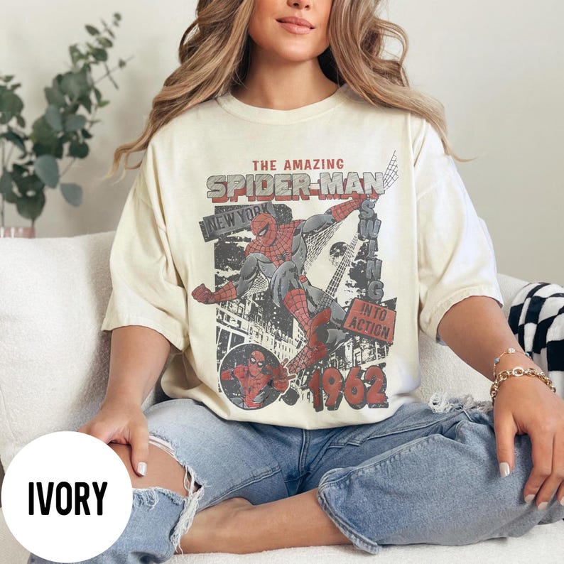 Pu&ograve; includere: T-shirt color avorio con grafica vintage di Spider-Man. Il design include il testo "THE AMAZING SPIDER-MAN NEW YORK" e "INTO ACTION 1962". La grafica raffigura Spider-Man in azione con uno sfondo di paesaggio urbano.