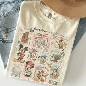 May include: Cream-colored t-shirt featuring a colorful graphic of various Disney characters and landmarks, including Mickey Mouse, Minnie Mouse, and Cinderella's castle. The shirt has the words "Walt Disney World" and the year "2026" printed on it.