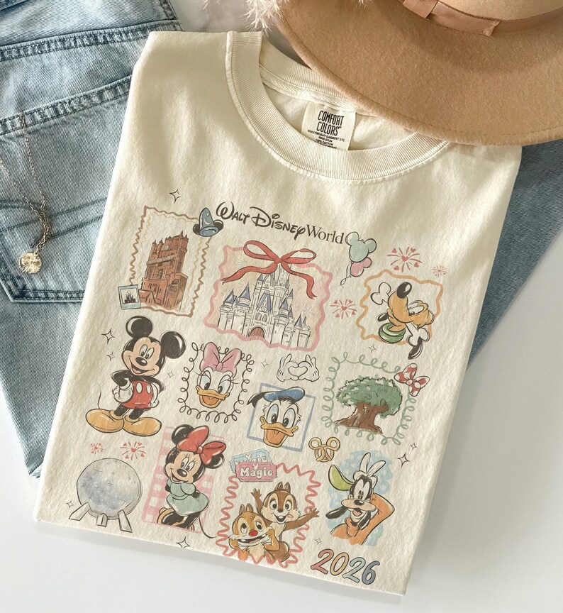 May include: Cream-colored t-shirt featuring a colorful graphic of various Disney characters and landmarks, including Mickey Mouse, Minnie Mouse, and Cinderella's castle. The shirt has the words "Walt Disney World" and the year "2026" printed on it.