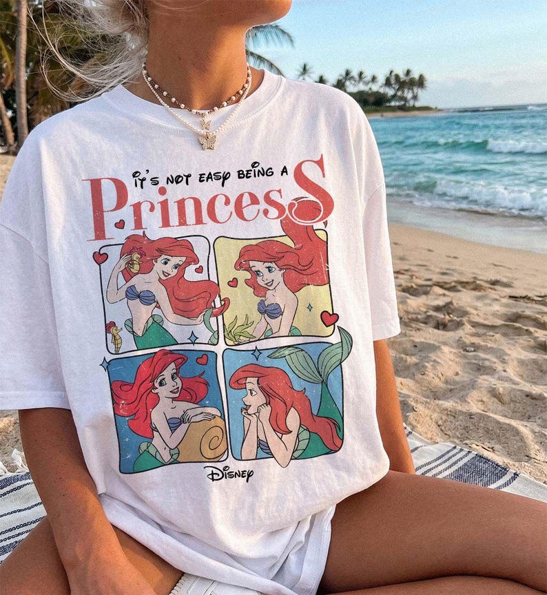 Puede incluir: Una camiseta blanca de manga corta con un estampado gr&aacute;fico del personaje Ariel de La Sirenita en cuatro paneles separados. El texto sobre los paneles dice It's not easy being a Princess, con el logotipo de Disney impreso en la parte inferior.