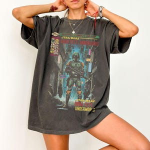 Puede incluir: Camiseta gris oscuro con un dise&ntilde;o de Boba Fett de Star Wars, en un callej&oacute;n de la ciudad. La camiseta incluye el texto "Marvel Comics Group Star Wars Boba Fett Underworld" en verde y rojo. El dise&ntilde;o es de inspiraci&oacute;n vintage.