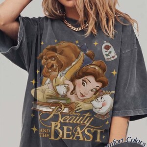 May include: A charcoal grey t-shirt featuring a graphic print of Belle, the Beast, Chip, and Mrs. Potts from Beauty and the Beast. The design includes a rose in a glass dome, yellow stars, and the text Beauty and the Beast. Comfort Colors Pepper.