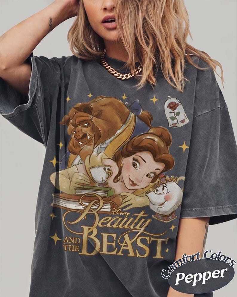 May include: A charcoal grey t-shirt featuring a graphic print of Belle, the Beast, Chip, and Mrs. Potts from Beauty and the Beast. The design includes a rose in a glass dome, yellow stars, and the text Beauty and the Beast. Comfort Colors Pepper.