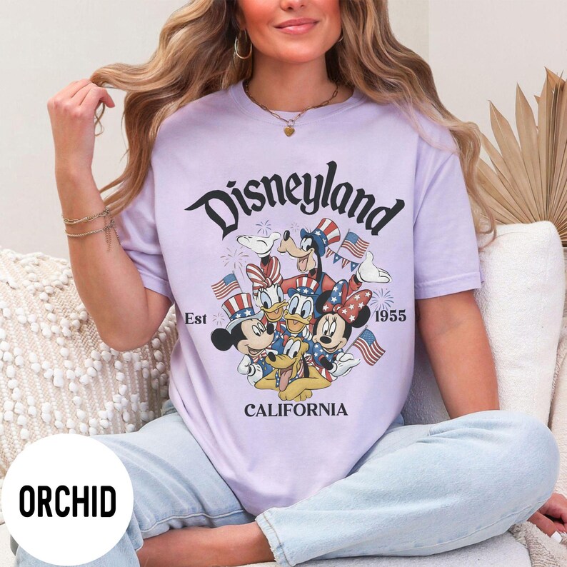 May include: Orchid-colored t-shirt featuring a Disneyland graphic with Mickey Mouse, Donald Duck, Goofy, and Minnie Mouse. The design includes American flags and the text "Disneyland California Est 1955."