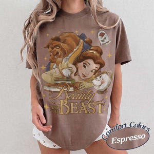 May include: An espresso-coloured t-shirt featuring a graphic print of Belle, the Beast, Chip, and Mrs. Potts from Beauty and the Beast. The design includes a rose in a glass dome, books, and yellow stars, with the text Beauty and the Beast printed below.