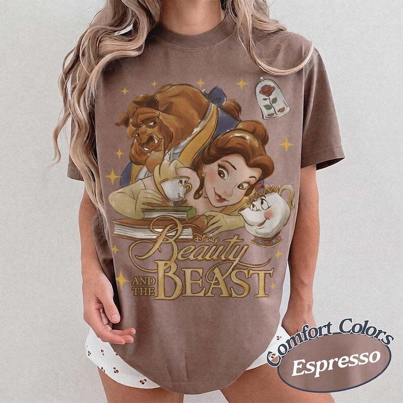 May include: An espresso-coloured t-shirt featuring a graphic print of Belle, the Beast, Chip, and Mrs. Potts from Beauty and the Beast. The design includes a rose in a glass dome, books, and yellow stars, with the text Beauty and the Beast printed below.