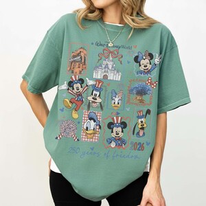 Może przedstawiać: Szałwiowo-zielony t-shirt z kr&oacute;tkim rękawem z nadrukiem postaci z Walt Disney World, w tym Myszki Miki, Minnie, Kaczora Donalda, Goofy'ego i Pluto w patriotycznych strojach. Wz&oacute;r zawiera tekst 250 years of freedom oraz 2026.