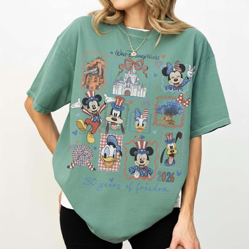 Może przedstawiać: Szałwiowo-zielony t-shirt z kr&oacute;tkim rękawem z nadrukiem postaci z Walt Disney World, w tym Myszki Miki, Minnie, Kaczora Donalda, Goofy'ego i Pluto w patriotycznych strojach. Wz&oacute;r zawiera tekst 250 years of freedom oraz 2026.