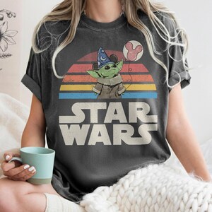 May include: Dark gray t-shirt featuring a graphic of a green alien character wearing a wizard hat and holding a Mickey Mouse balloon. The Star Wars logo is below a retro sunset design.