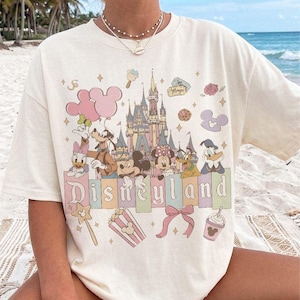 May include: A cream-coloured short-sleeve t-shirt featuring a graphic print of the Disneyland castle, Mickey Mouse, Minnie Mouse, Goofy, Donald Duck, and Daisy Duck. The design includes pastel-coloured balloons, a bow, popcorn, and a drink.
