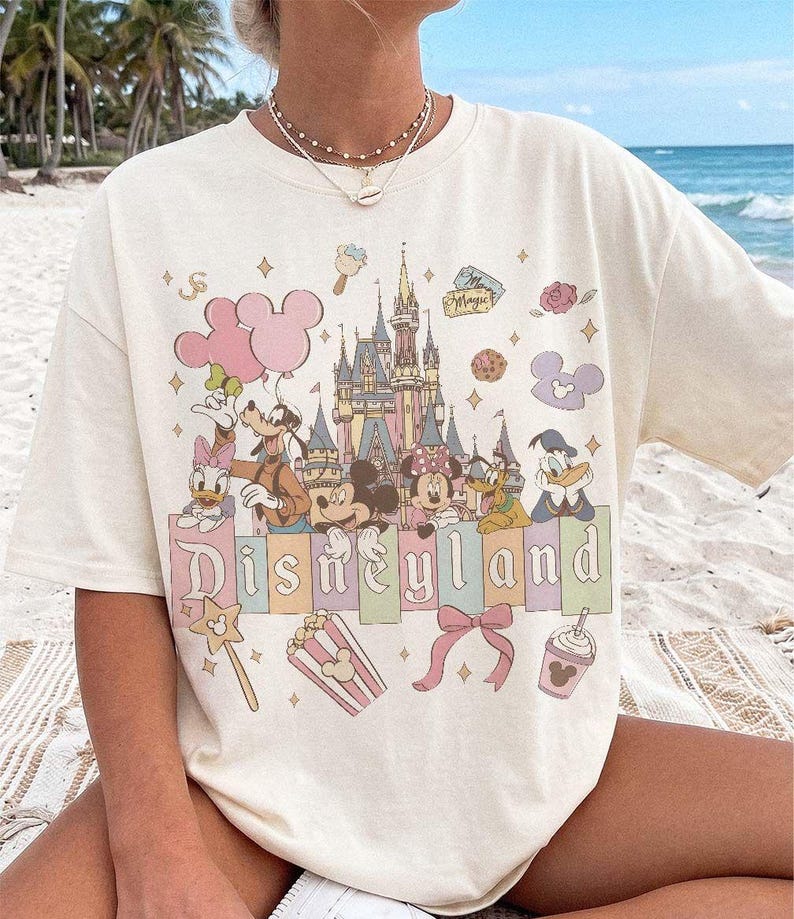 May include: A cream-coloured short-sleeve t-shirt featuring a graphic print of the Disneyland castle, Mickey Mouse, Minnie Mouse, Goofy, Donald Duck, and Daisy Duck. The design includes pastel-coloured balloons, a bow, popcorn, and a drink.
