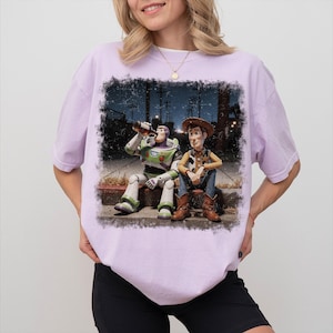 May include: A light purple short-sleeve t-shirt featuring a graphic print of the characters Buzz Lightyear and Woody sitting on a curb at night. Buzz Lightyear is drinking from a brown bottle, while Woody sits beside him in a cowboy outfit.