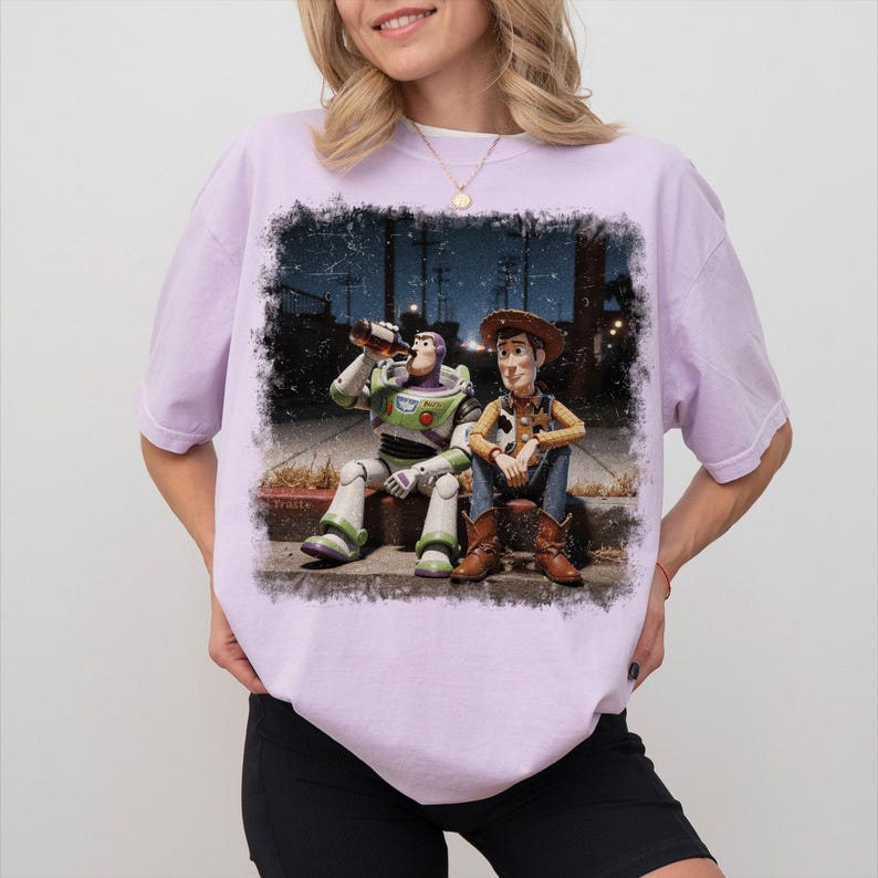 May include: A light purple short-sleeve t-shirt featuring a graphic print of the characters Buzz Lightyear and Woody sitting on a curb at night. Buzz Lightyear is drinking from a brown bottle, while Woody sits beside him in a cowboy outfit.