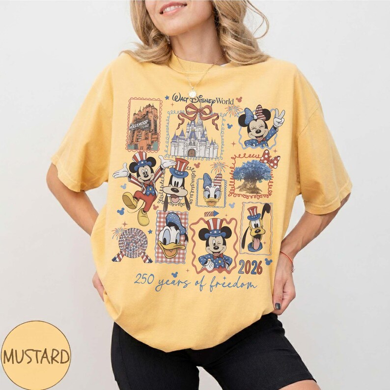 Może przedstawiać: Musztardowy t-shirt z kr&oacute;tkim rękawem z nadrukiem graficznym przedstawiającym postacie Disneya w patriotycznych strojach, w tym Myszkę Miki, Myszkę Minnie, Goofy'ego i Kaczora Donalda, z napisem Walt Disney World, 2026 oraz 250 years of freedom.