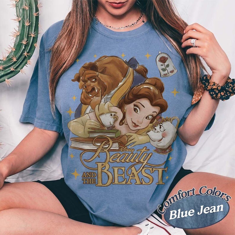 May include: A blue t-shirt featuring a graphic print of Belle, the Beast, Chip, and Mrs. Potts from Beauty and the Beast. The design includes a rose in a glass dome, books, and gold stars, with the text Beauty and the Beast printed in gold.