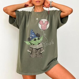 K&ouml;nnte beinhalten: Olivgr&uuml;nes T-Shirt mit einer Grafik von Yoda mit Zauberhut und Mickey Mouse-Ballon. Das Wort "Yoda" ist in einer verspielten Schrift geschrieben. Das T-Shirt hat kurze &Auml;rmel und eine lockere Passform.