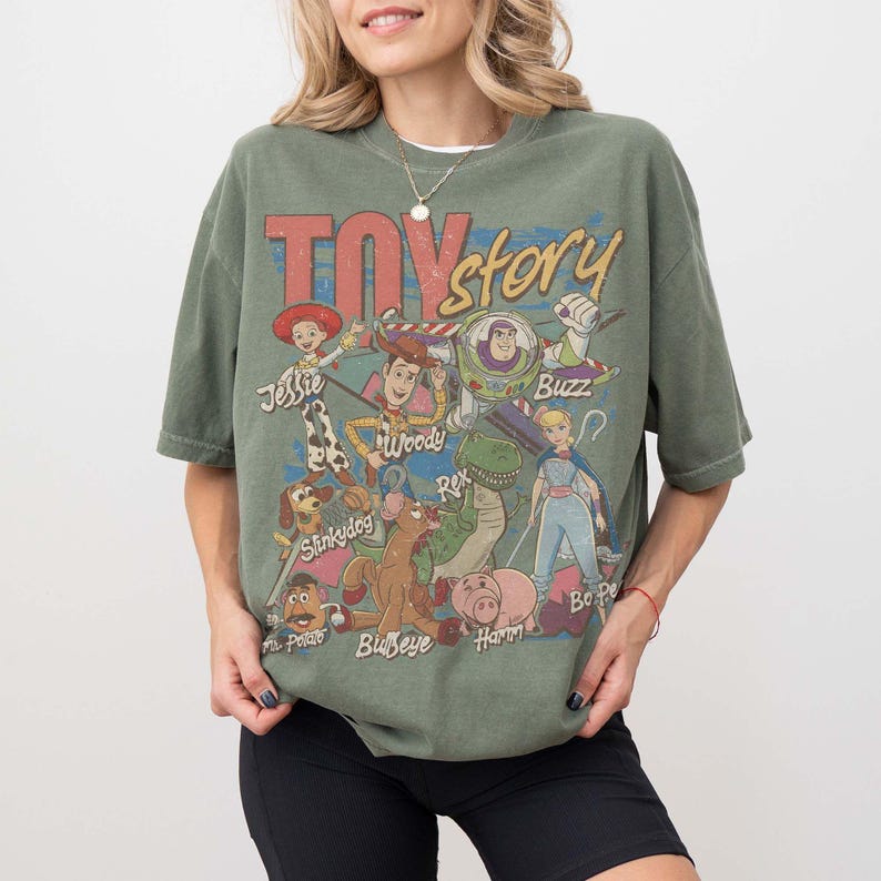 May include: An olive green short-sleeve t-shirt featuring a distressed graphic print of Toy Story characters including Woody, Buzz Lightyear, Jessie, Rex, Slinky Dog, Bullseye, Hamm, Mr. Potato Head, and Bo Peep with their names printed below.