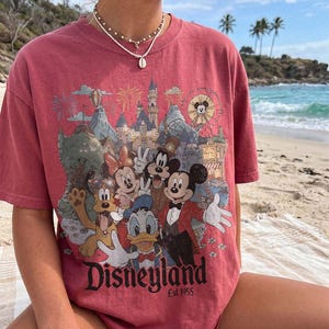 May include: A faded red short-sleeve t-shirt featuring a graphic print of Mickey Mouse, Minnie Mouse, Donald Duck, Goofy, and Pluto in front of a castle. The text Disneyland Est. 1955 is printed at the bottom of the graphic in a black serif font.