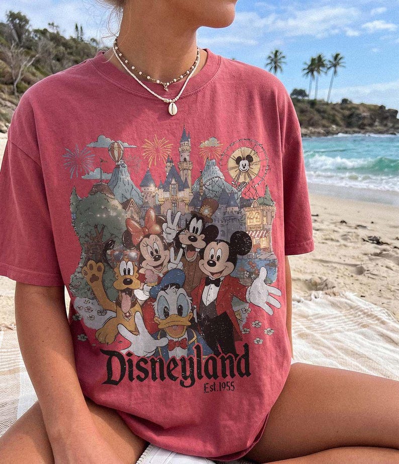 May include: A faded red short-sleeve t-shirt featuring a graphic print of Mickey Mouse, Minnie Mouse, Donald Duck, Goofy, and Pluto in front of a castle. The text Disneyland Est. 1955 is printed at the bottom of the graphic in a black serif font.