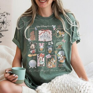 May include: Green t-shirt featuring a collage of Disney characters and landmarks, including Mickey Mouse, Minnie Mouse, and Cinderella's castle. The shirt has the text "Walt Disney World" and "The Most Magical Place on Earth" with the year "2026".