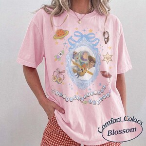 Peut inclure: Un t-shirt rose clair &agrave; manches courtes orn&eacute; d'un cadre bleu ornement&eacute; contenant une illustration de Buzz l'&Eacute;clair et Woody. Autour du cadre se trouvent des motifs de plan&egrave;te, de fus&eacute;e, d'&eacute;toiles et le texte TO INFINITY AND BEYOND.