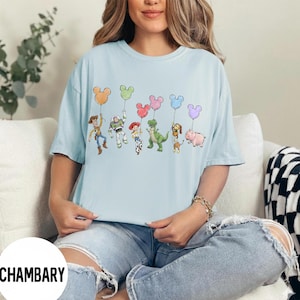 K&ouml;nnte beinhalten: Hellblaues T-Shirt mit einer bunten Grafik von Zeichentrickfiguren, die Mickey Mouse-f&ouml;rmige Ballons halten. Zu den Figuren geh&ouml;ren ein Cowboy, ein Weltraumranger, ein Cowgirl, ein gr&uuml;ner Dinosaurier, ein Hund und ein Schwein. Das Shirt ist ein l&auml;ssiger Stil mit kurzen &Auml;rmeln.