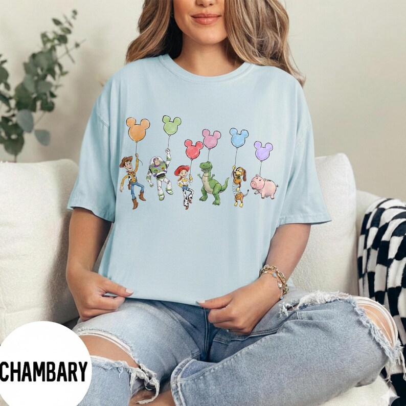K&ouml;nnte beinhalten: Hellblaues T-Shirt mit einer bunten Grafik von Zeichentrickfiguren, die Mickey Mouse-f&ouml;rmige Ballons halten. Zu den Figuren geh&ouml;ren ein Cowboy, ein Weltraumranger, ein Cowgirl, ein gr&uuml;ner Dinosaurier, ein Hund und ein Schwein. Das Shirt ist ein l&auml;ssiger Stil mit kurzen &Auml;rmeln.