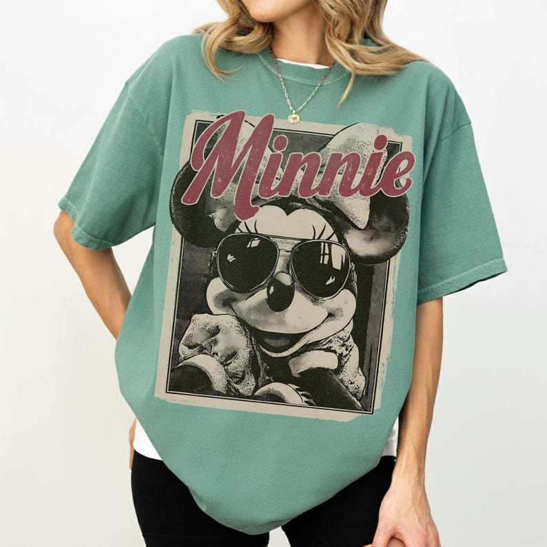 May include: A sage green short-sleeve t-shirt featuring a graphic print of Minnie Mouse wearing dark sunglasses. The word Minnie is printed in a large, distressed maroon font above the character. The design is framed within a rectangular border.