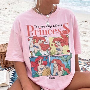 Puede incluir: Una camiseta rosa claro con un estampado gr&aacute;fico del personaje Ariel de La Sirenita en cuatro paneles distintos. El texto dice It's not easy being a Princess y Disney. La camiseta tiene cuello redondo y mangas cortas.