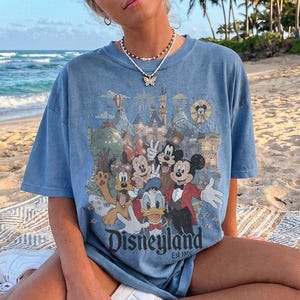 May include: A blue short-sleeve t-shirt featuring a graphic print of Mickey Mouse, Minnie Mouse, Donald Duck, Goofy, and Pluto in front of a castle. The text Disneyland Est. 1955 is printed at the bottom of the graphic in a dark blue font.
