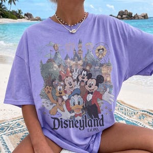 May include: A light purple short-sleeve t-shirt featuring a graphic print of Mickey Mouse, Minnie Mouse, Donald Duck, Goofy, and Pluto in front of a castle. The text "Disneyland Est. 1955" is printed below the characters in a dark font.