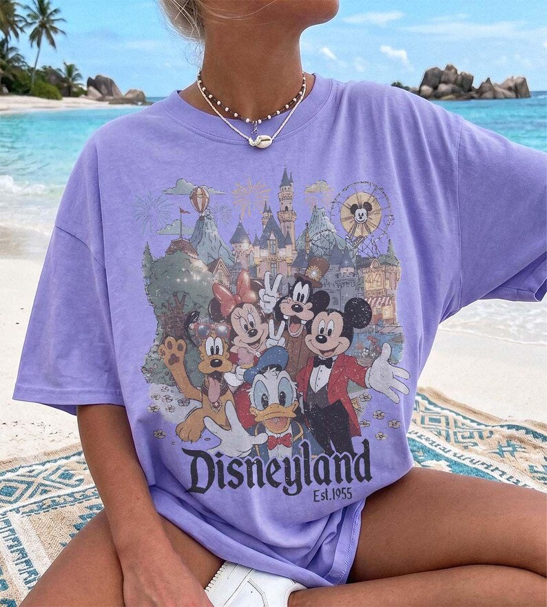 May include: A light purple short-sleeve t-shirt featuring a graphic print of Mickey Mouse, Minnie Mouse, Donald Duck, Goofy, and Pluto in front of a castle. The text "Disneyland Est. 1955" is printed below the characters in a dark font.