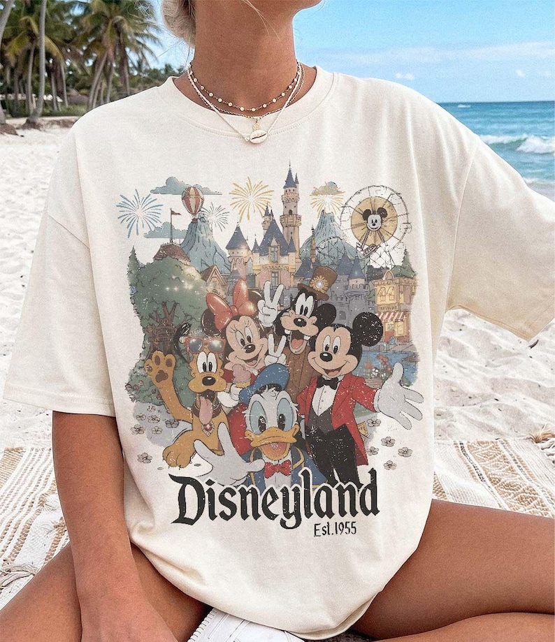May include: A cream-colored short-sleeve t-shirt featuring a graphic print of Mickey Mouse, Minnie Mouse, Donald Duck, Goofy, and Pluto in front of a castle. The text "Disneyland Est. 1955" is printed in black at the bottom of the graphic design.