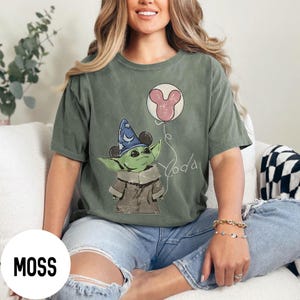 K&ouml;nnte beinhalten: Moosgr&uuml;nes T-Shirt mit einer Grafik eines gr&uuml;nen Alien-Charakters, der einen Zaubererhut tr&auml;gt und einen Mickey-Mouse-Ballon h&auml;lt. Das Wort "Yoda" ist in Schreibschrift geschrieben. Das Shirt hat eine lockere Passform.