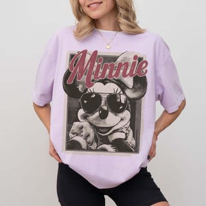 May include: A light purple t-shirt featuring a graphic print of Minnie Mouse wearing sunglasses. The design includes the text "Minnie" in a large, distressed red font above the character, set within a rectangular frame on the front of the shirt.