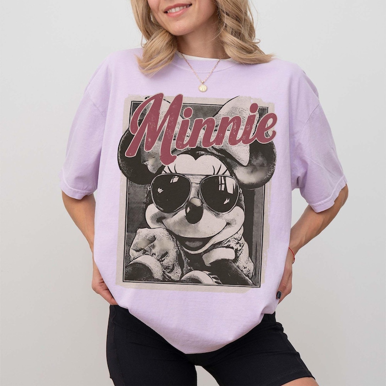 May include: A light purple t-shirt featuring a graphic print of Minnie Mouse wearing sunglasses. The design includes the text "Minnie" in a large, distressed red font above the character, set within a rectangular frame on the front of the shirt.