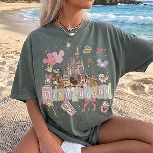May include: A sage green short-sleeve t-shirt featuring a graphic print of a castle, Mickey Mouse, Minnie Mouse, Goofy, Donald Duck, and Daisy Duck. The text Disneyland is printed across the centre, surrounded by icons of balloons, popcorn, and a bow.