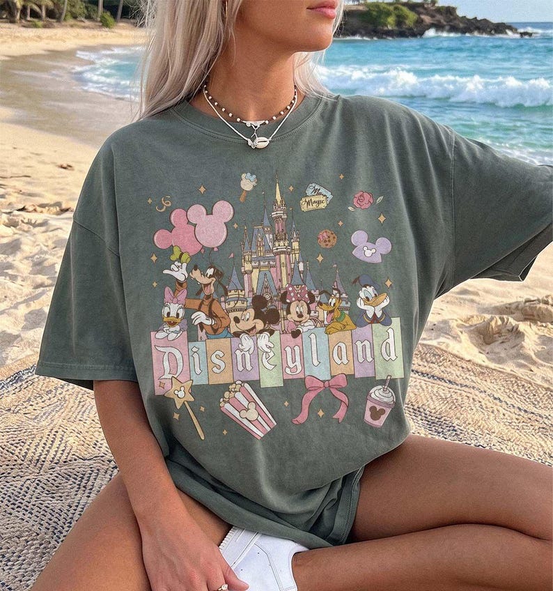 May include: A sage green short-sleeve t-shirt featuring a graphic print of a castle, Mickey Mouse, Minnie Mouse, Goofy, Donald Duck, and Daisy Duck. The text Disneyland is printed across the centre, surrounded by icons of balloons, popcorn, and a bow.
