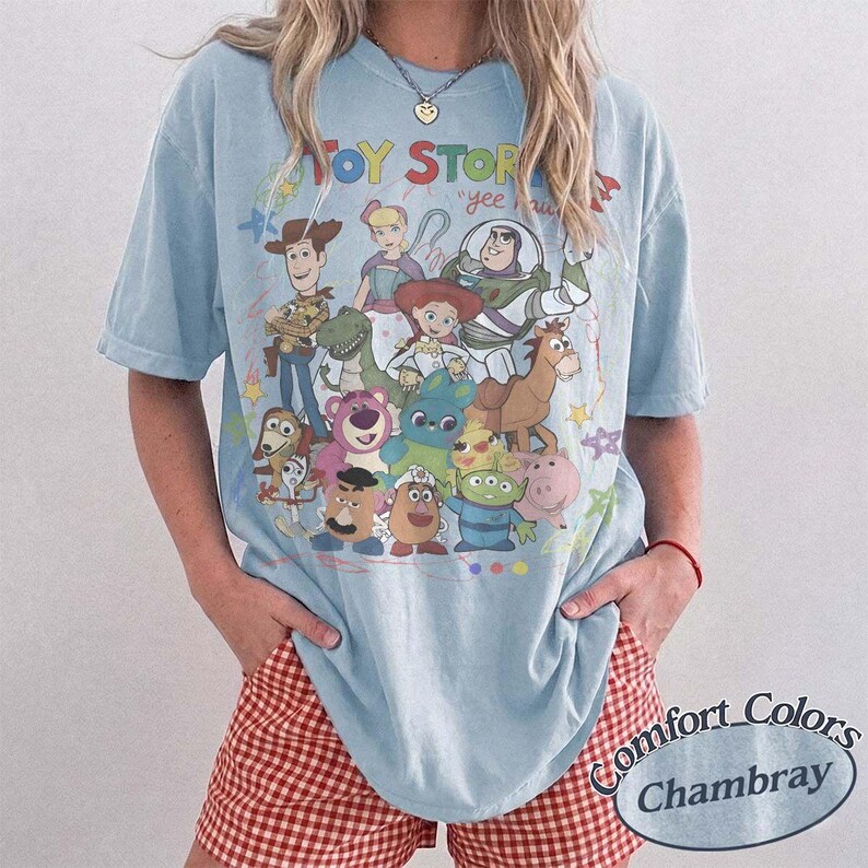 May include: A light blue short-sleeve t-shirt featuring a colorful graphic print of various Toy Story characters including Woody, Buzz Lightyear, Jessie, and Rex. The text "TOY STORY" is printed in bold letters above the character illustration.
